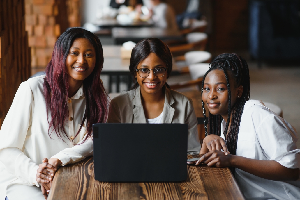 Three African girls (students) sitting at the table in cafe studying up for test or making homework together, they are using laptop and digital tablet.